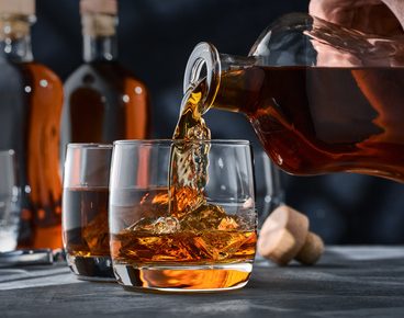 Two glasses of whiskey with ice on a concrete table, a glass of whiskey is poured from a bottle. Two glasses of brandy on a concrete table, brandy from a bottle being poured into the glass.