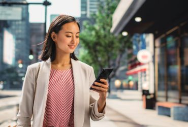 Portrait of an Attractive Japanese Female Wearing Smart Casual Clothes and Using Smartphone on the Urban Street. Manager in Big City Connecting with People Online, Messaging and Browsing Internet.