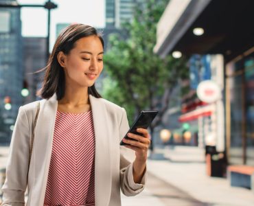 Portrait of an Attractive Japanese Female Wearing Smart Casual Clothes and Using Smartphone on the Urban Street. Manager in Big City Connecting with People Online, Messaging and Browsing Internet.