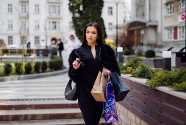 A brown-haired woman wearing black wear, holds colourful, patterned shopping bags during a successful shopping spree. Walking outside, she is enjoying the warmth of a day A brown-haired woman wearing black wear, holds colourful, patterned shopping bags during a successful shopping spree. Walking outside, she is enjoying the warmth of a day