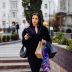 A brown-haired woman wearing black wear,  holds colourful, patterned shopping bags during a successful shopping spree. Walking outside, she is enjoying the warmth of a day